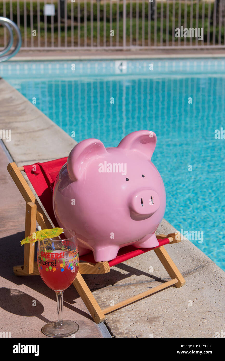 Pink piggy bank on a red deck chair relaxing by a pool on a hot summer day with a glass marked 'cheers'!  and cocktail umbrella Stock Photo