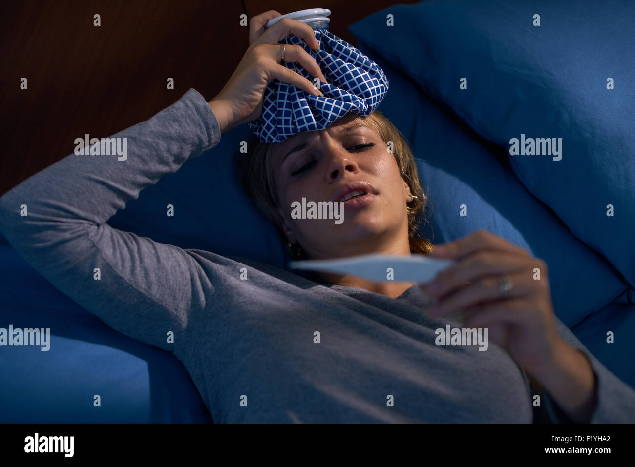 Young hispanic woman in pajamas, laying on bed with fever and ice bag on head, measuring temperature with digital thermometer at Stock Photo