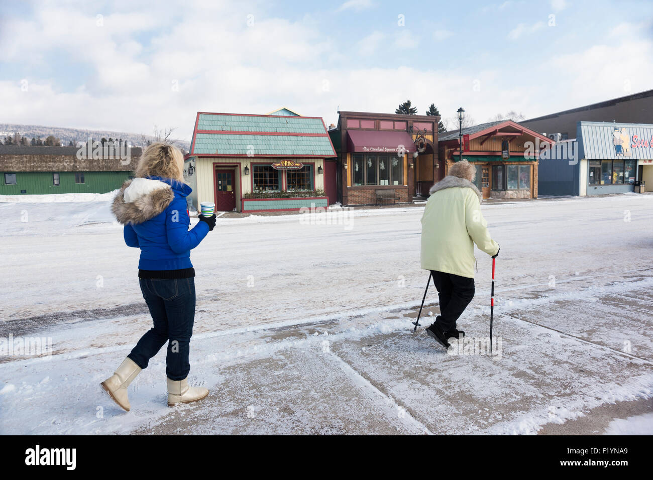 Two Caucasian woman wearing coats and boots walk along the sidewalk past shops in town during winter, Grand Marais, MN, USA Stock Photo