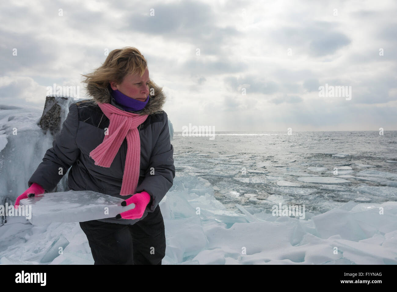Blond Caucasian woman in her 40s wearing a winter coat and pink scarf prepares to throw a large chunk of plate ice on the shore Stock Photo