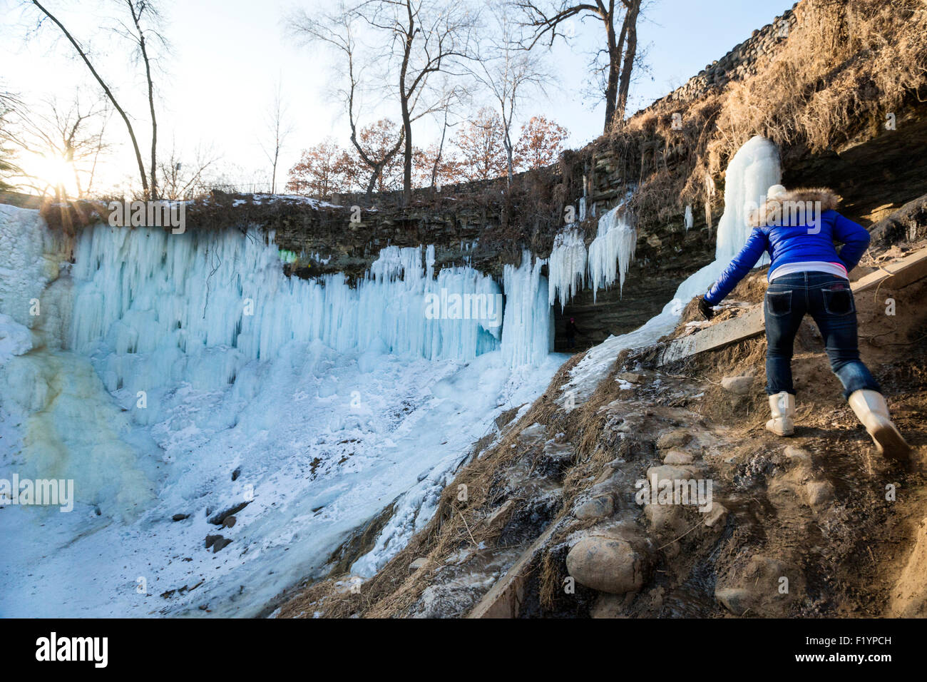 A woman in coat, gloves and boots hikes up slippery rocks to reach frozen Minnehaha Falls in winter, Minneapolis, MN, USA Stock Photo