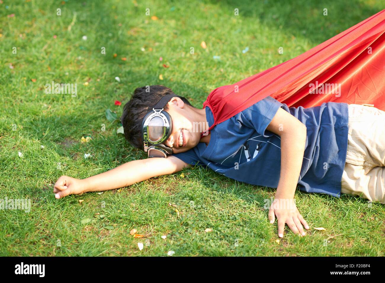 Boy wearing goggles and cape lying on side on grass, flying stance Stock Photo