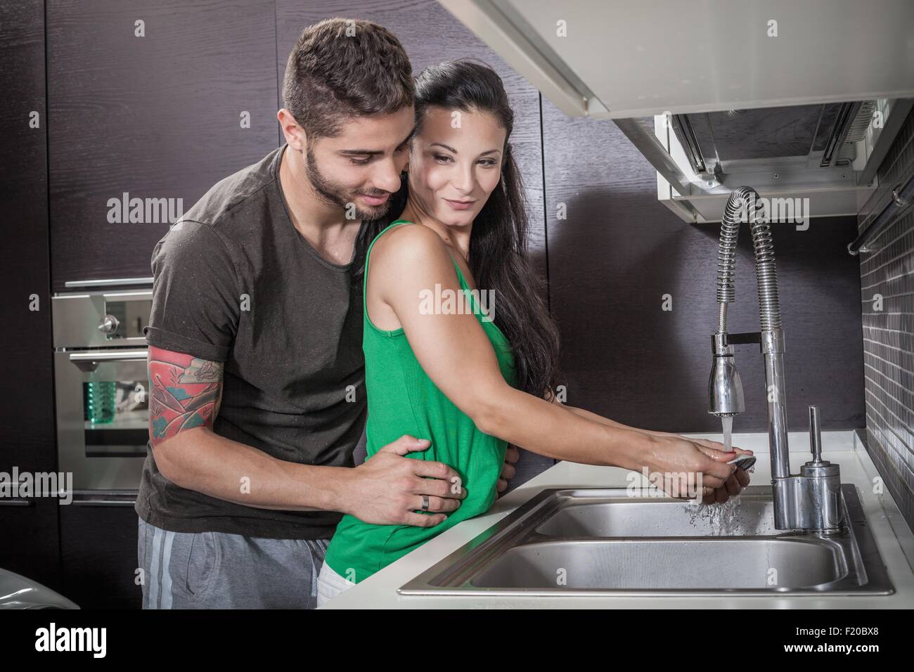 Young woman washing up whilst being embraced by boyfriend Stock Photo