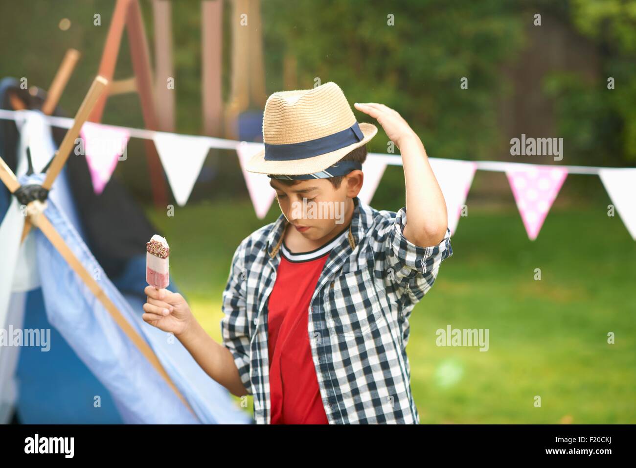 Boy eating ice lolly whilst adjusting trilby hat in garden Stock Photo