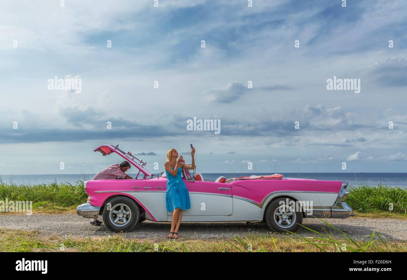 Young man trying to fix a vintage convertible at coast whilst girlfriend putting on make up, Havana, Cuba Stock Photo