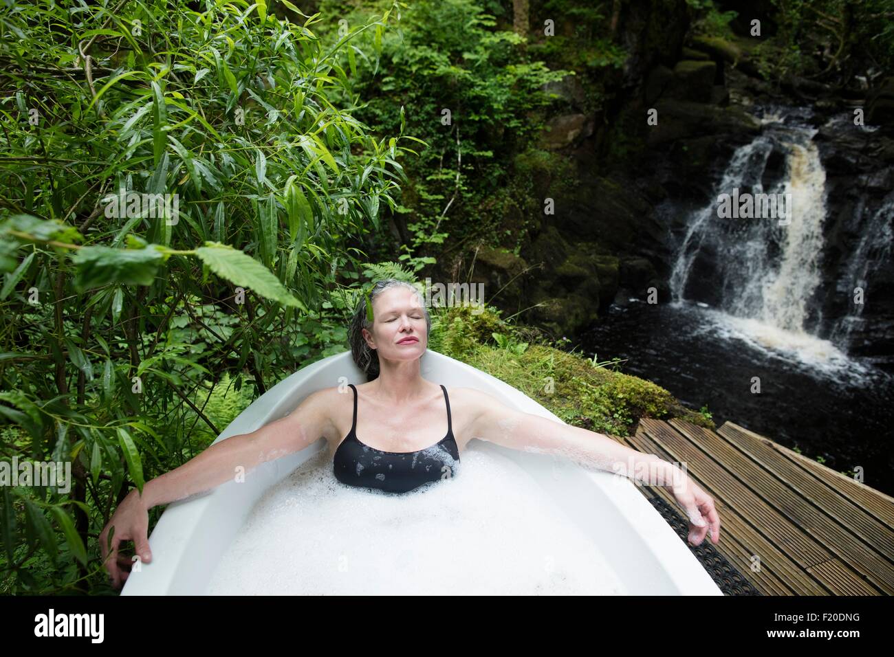 Mature woman relaxing in bubble bath in front of waterfall at eco retreat Stock Photo