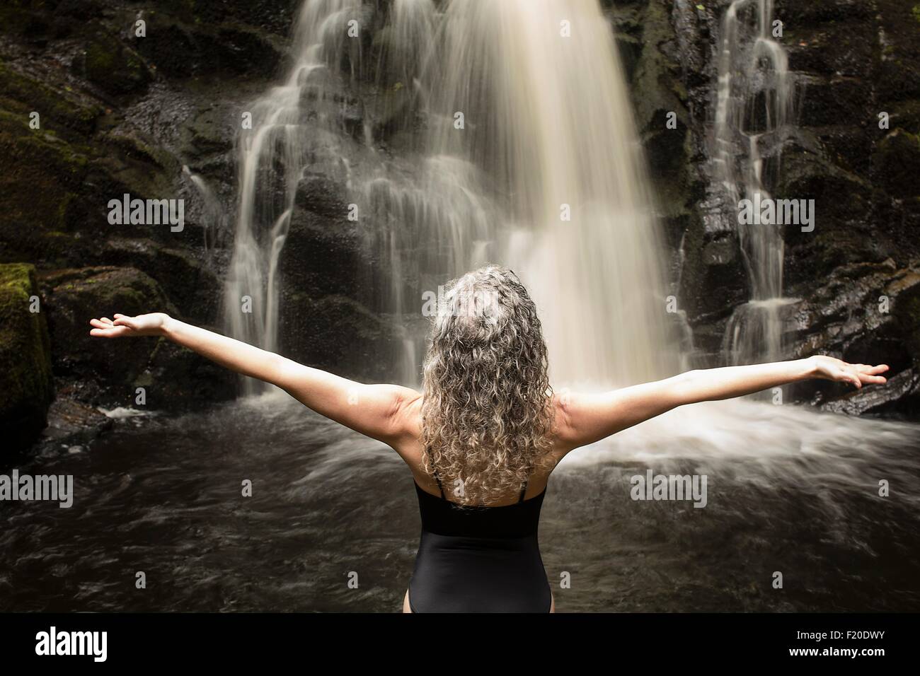 Rear view of mature woman practicing yoga pose in front of waterfall ...