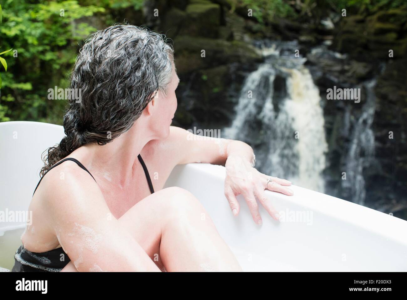 Mature woman in bubble bath gazing at waterfall at eco retreat Stock Photo