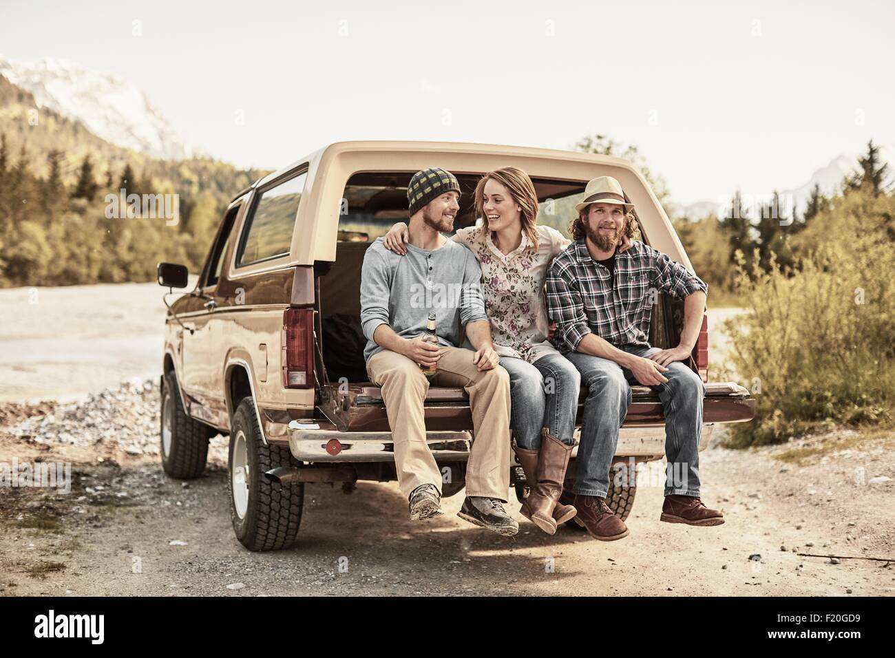 Three people sitting on back of pickup truck, arms around shoulders, smiling Stock Photo