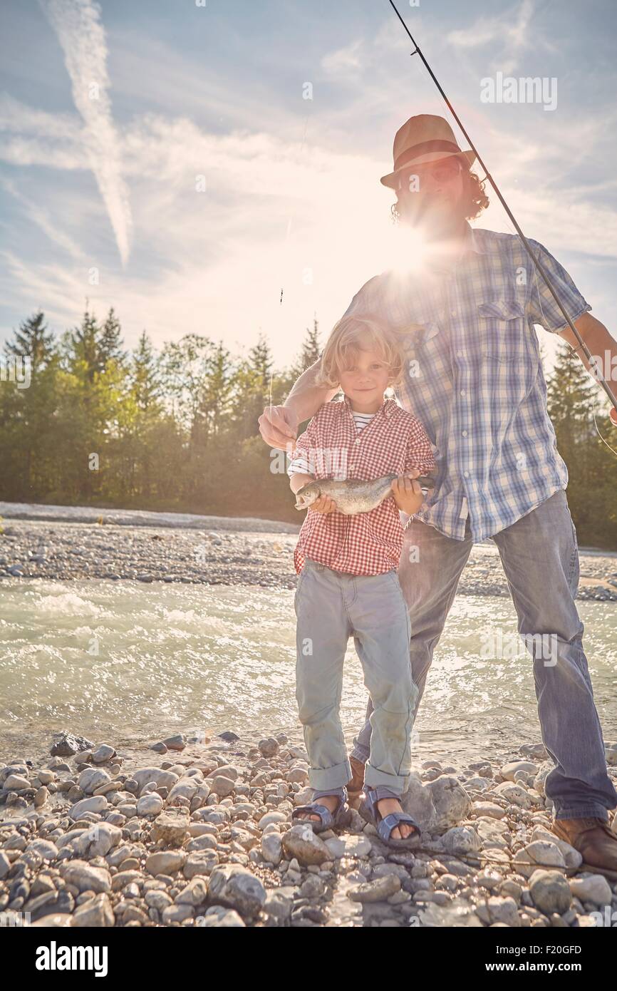 Mid adult man and boy next to river standing showing off fish, looking at camera Stock Photo