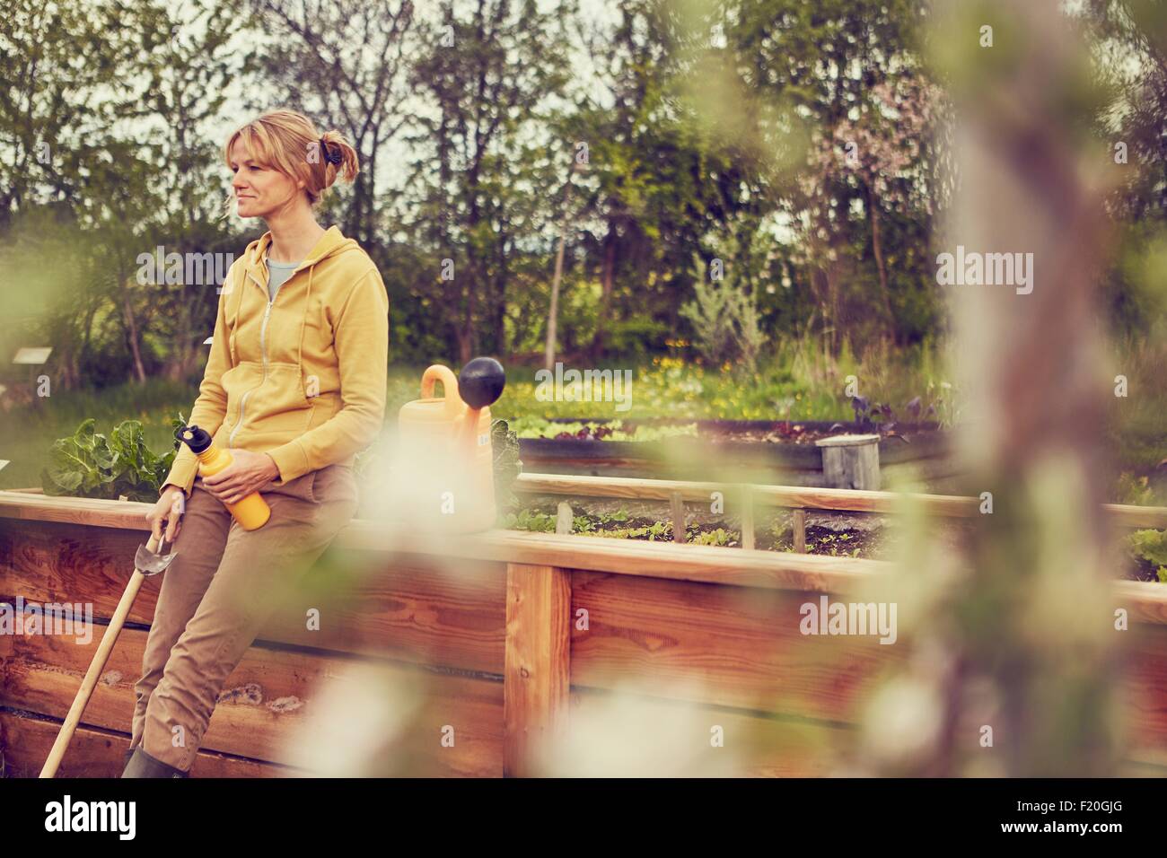 Mature woman, leaning against fence, taking a break from gardening, holding trowel and water bottle Stock Photo