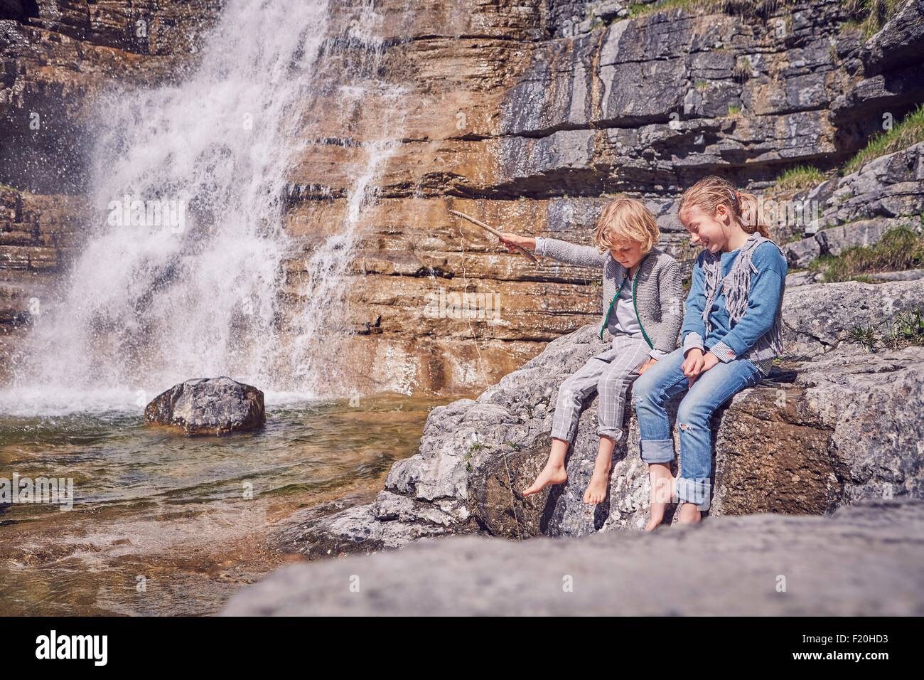 Brother and sister, sitting on rock, relaxing, beside waterfall Stock Photo