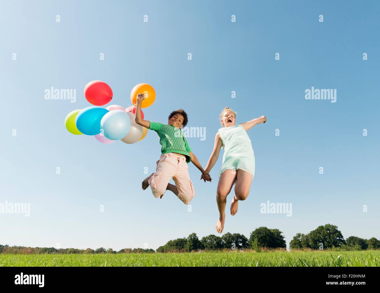 Girl and boy holding bunch of balloons jumping mid air in field Stock Photo