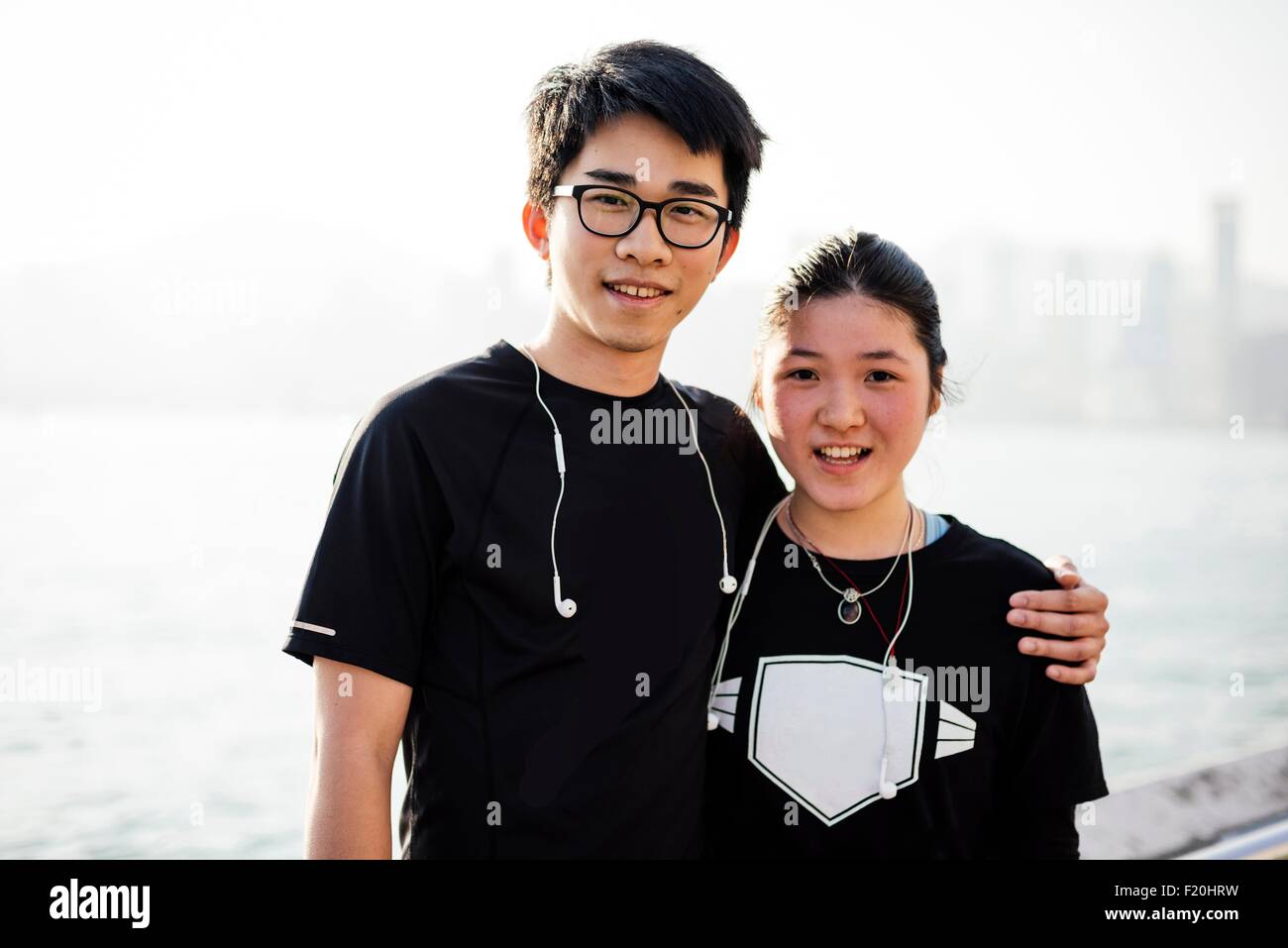 Portrait of young man with arm around young woman, wearing earphones, looking at camera smiling Stock Photo