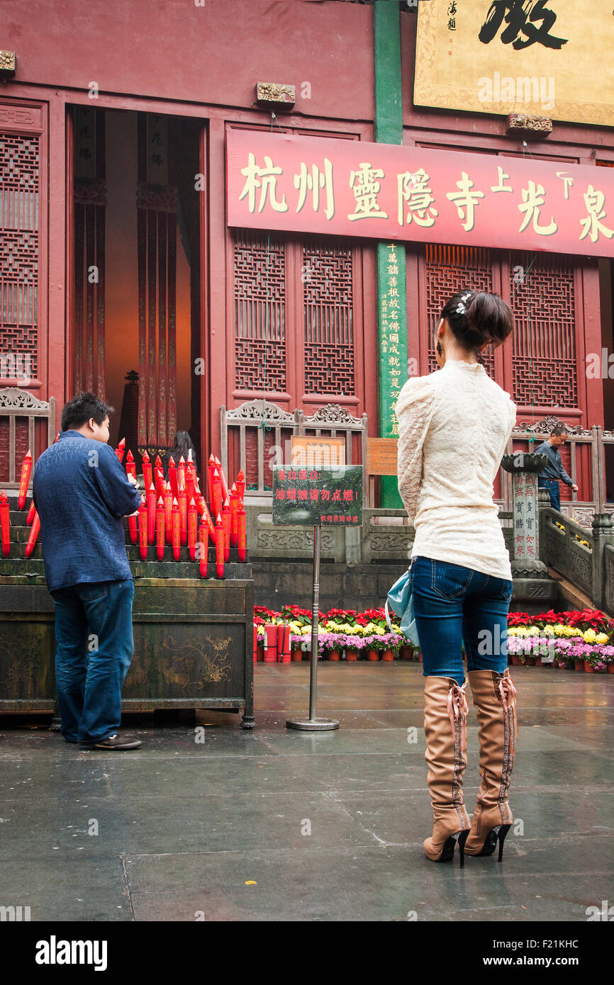 Asian woman in tall boots watches a man light candles while  worshiping at Lingyin Temple, oldest Buddhist temple in China Stock Photo