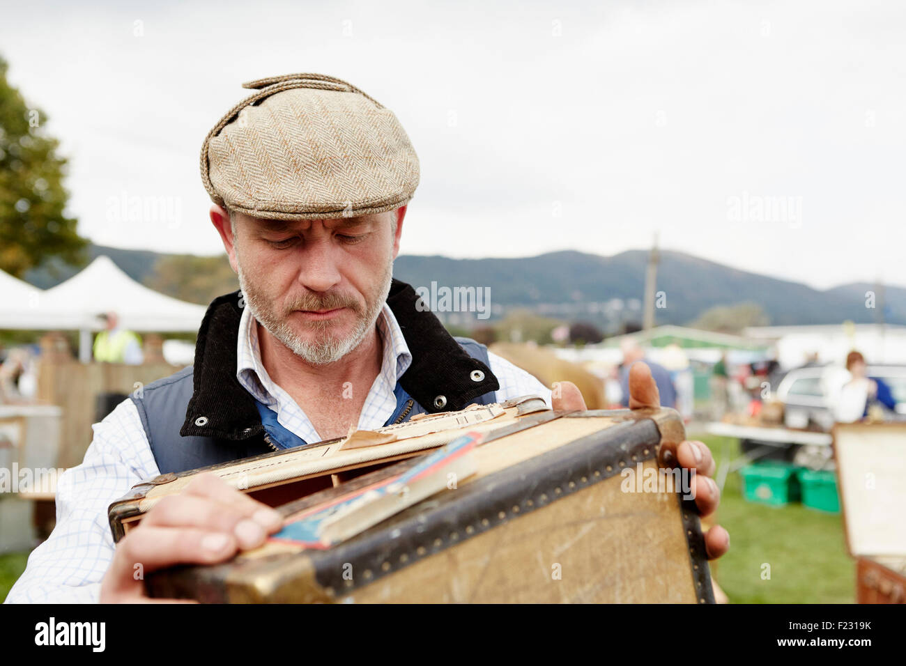 Man wearing a flat cap looking at a vintage suitcase at a flea market. Stock Photo