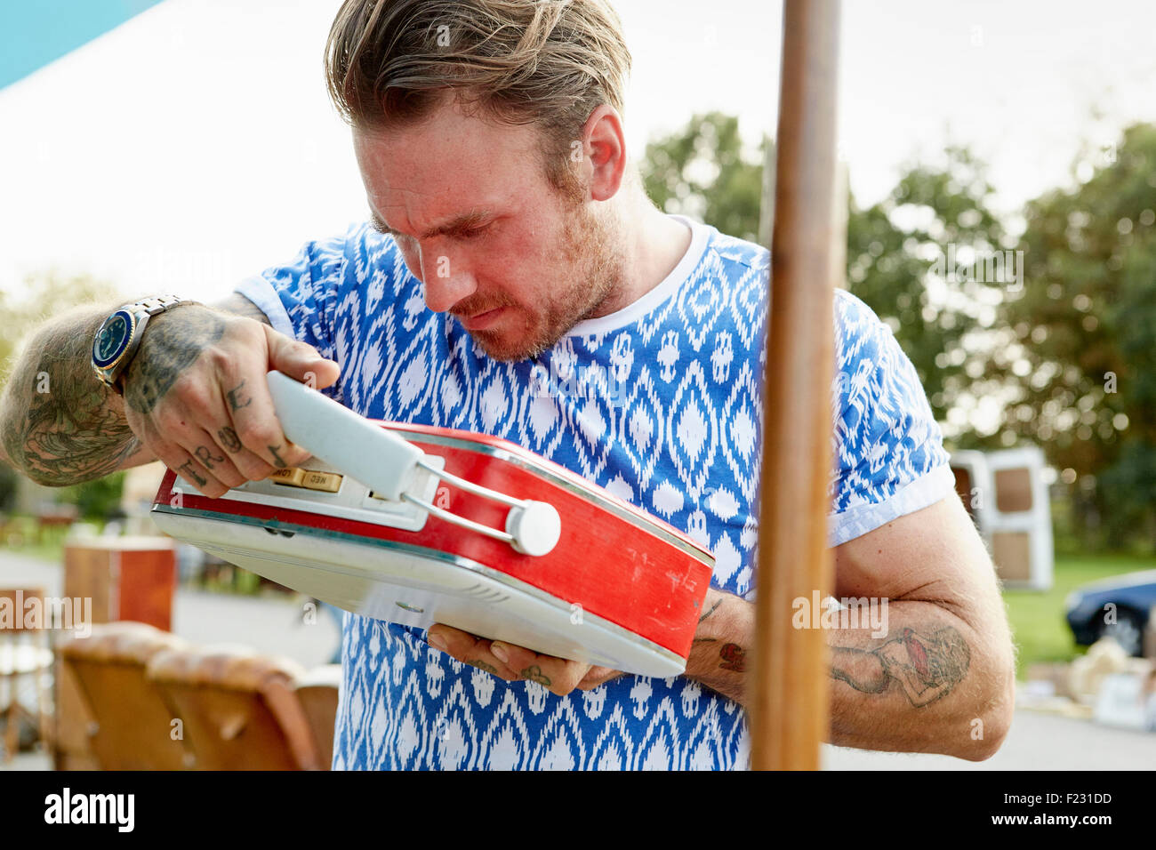 A man in a blue shirt with tattooed forearms, looking at a red vintage radio at a flea market. Stock Photo