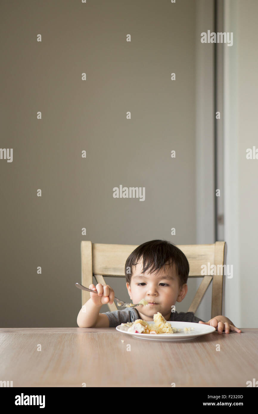 Young boy sitting on a chair at a table, eating a slice of cake. Stock Photo