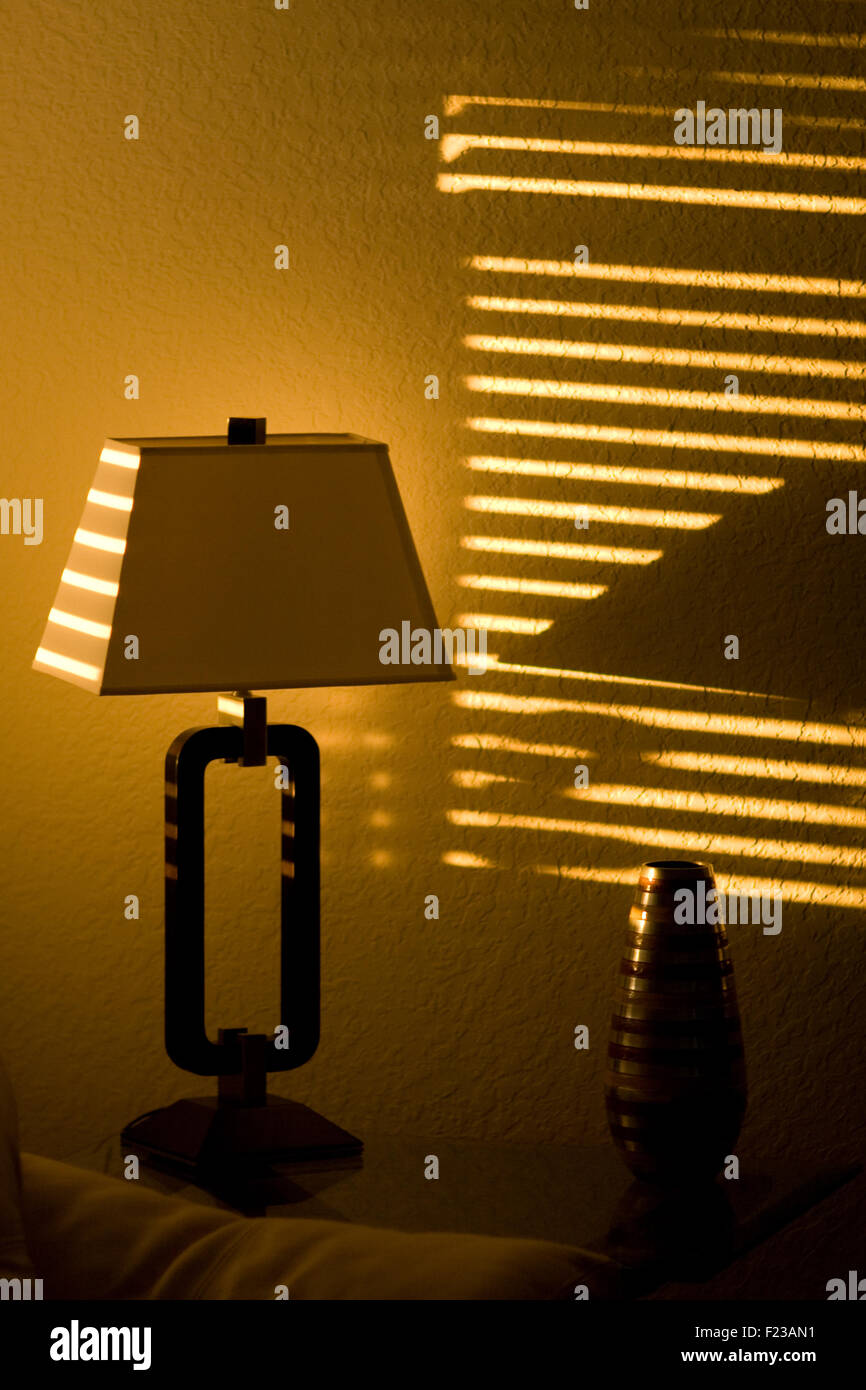 Bright sunshine in a dark room casts lines through window blinds and on to a lamp. Stock Photo