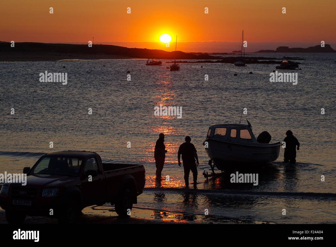 Sunset and boats moored in the bay, people bringing moter boat out of water to trailer on beach Stock Photo