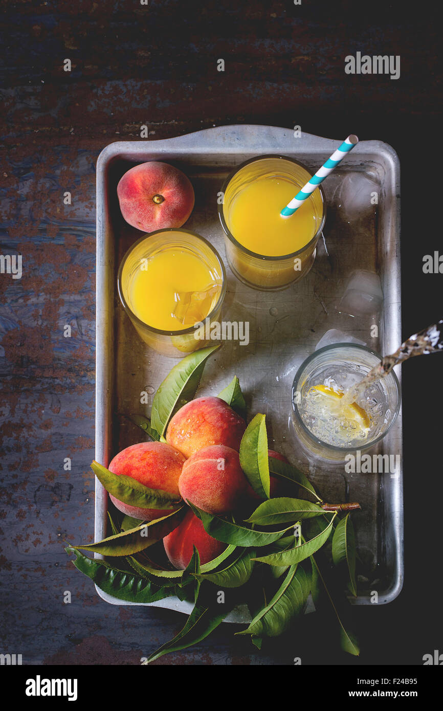 Peaches on branch with leaves and glasses with peach juice and limonade with ice cubes in aluminum tray over old metal table. Da Stock Photo