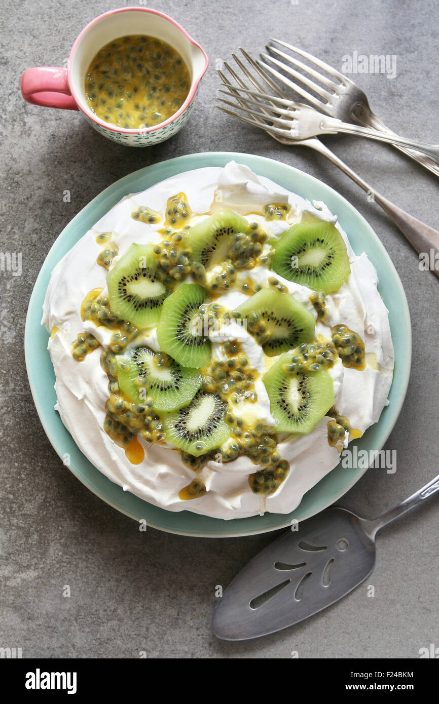 Pavlova with kiwi and passion fruit on a plate with a jug of passion fruit juice and cutlery over grey background.Top view Stock Photo