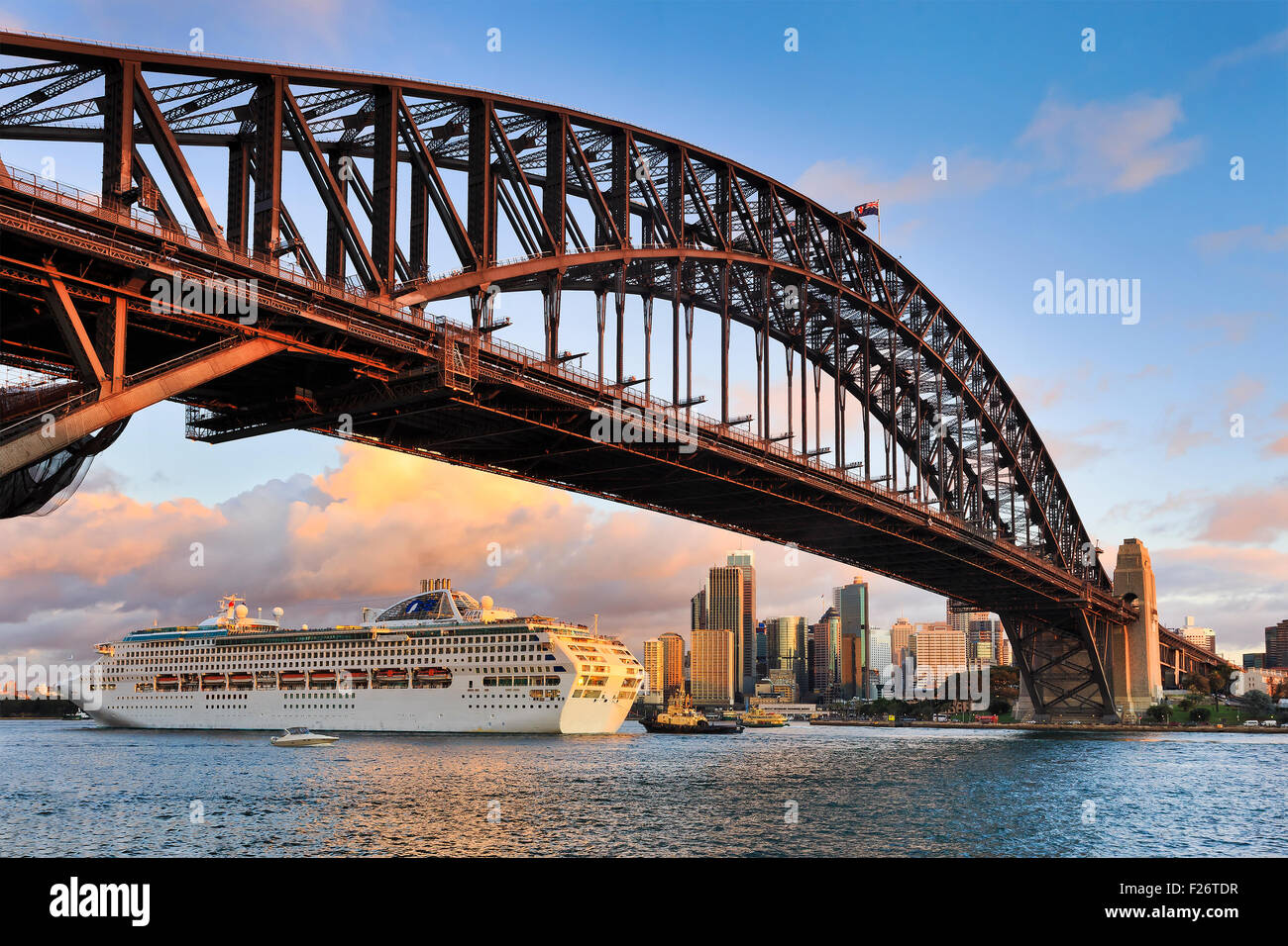 Oversized ocean cruise liner passing by under the Sydney Harbour Bridge at sunset backgrounded by city skyscrapers Stock Photo