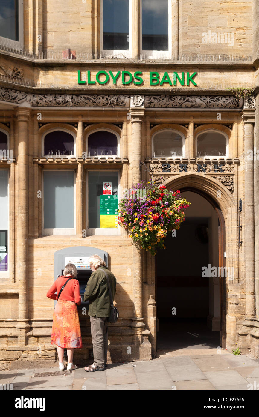 A couple using the ATM cash machine outside Lloyds bank; Glastonbury branch, High St, Glastonbury town, Somerset England UK Stock Photo
