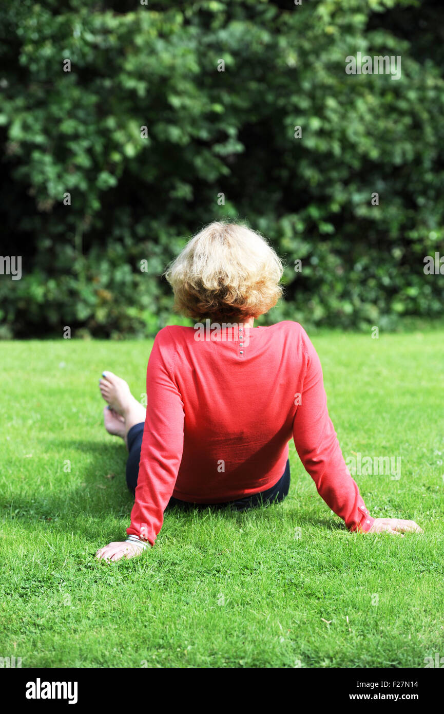 Back view of middle aged woman wearing coral pink coloured top and blue trousers sitting on grass outdoors Stock Photo