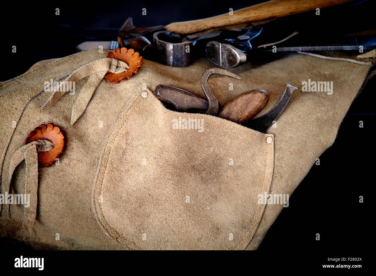 Tools of a farrier including rasp, hoof knife, chaps, hoof pick, and nippers draped over stool Stock Photo