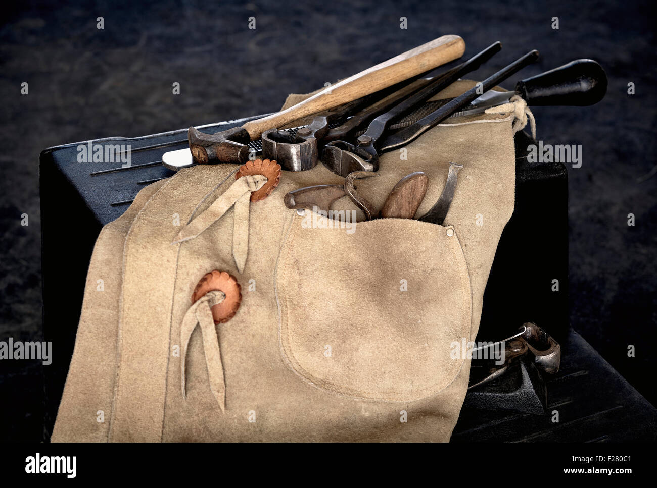 Tools of a farrier including rasp, hoof knife, chaps, hoof pick, and nippers draped over stool Stock Photo