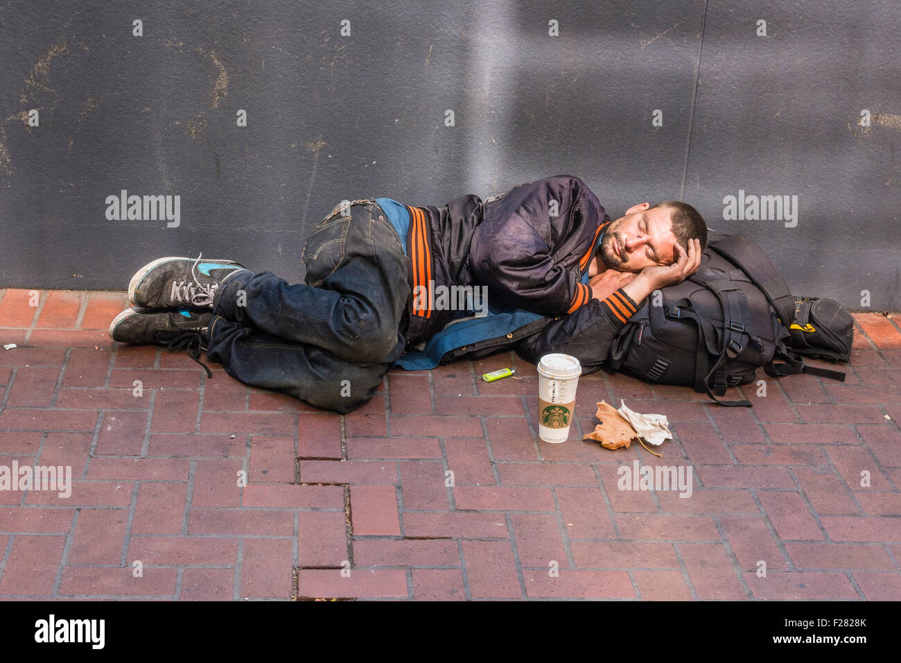 A 30s homeless man sleeps, lying down, on a sidewalk in San Francisco. He wears a Giants major league baseball jacket. Stock Photo