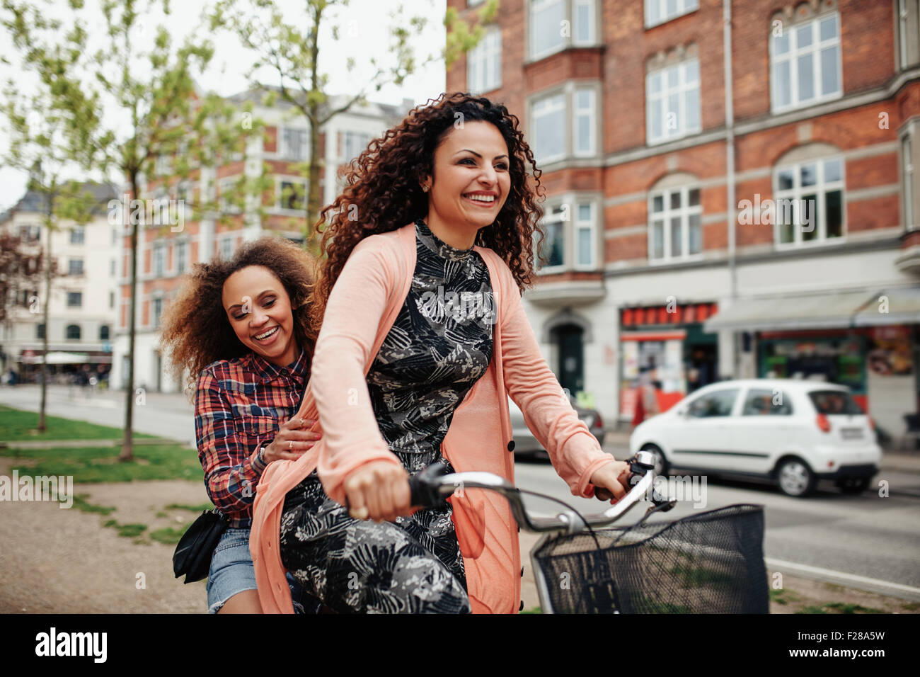 Portrait of cheerful young friends riding a bicycle in the city. Young girls outdoors enjoying bicycle ride on street. Stock Photo