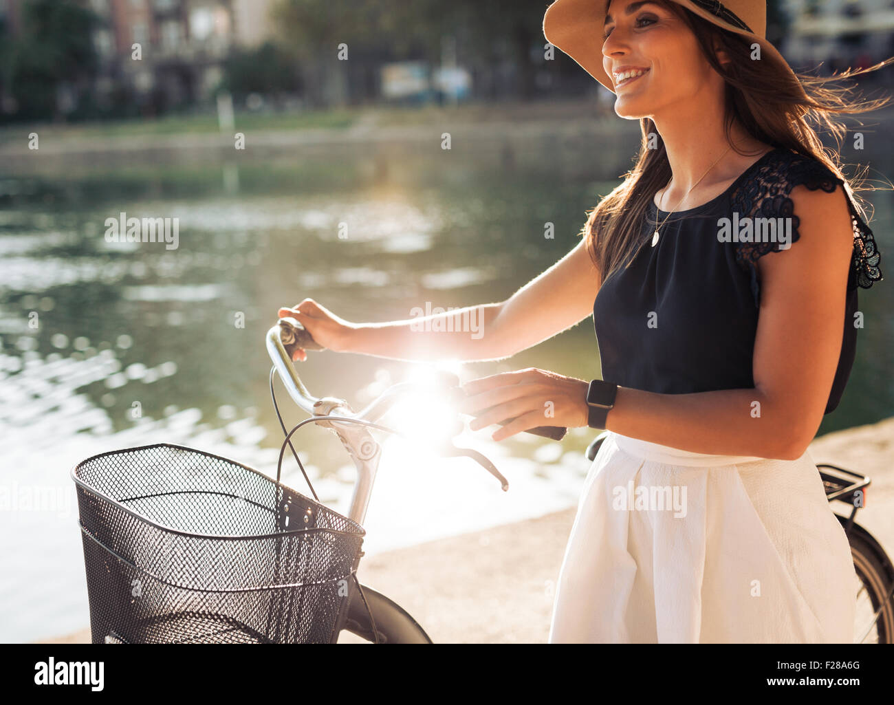 Cheerful young female at the park with her bicycle. Female model wearing hat looking away smiling while walking along a pond. Stock Photo