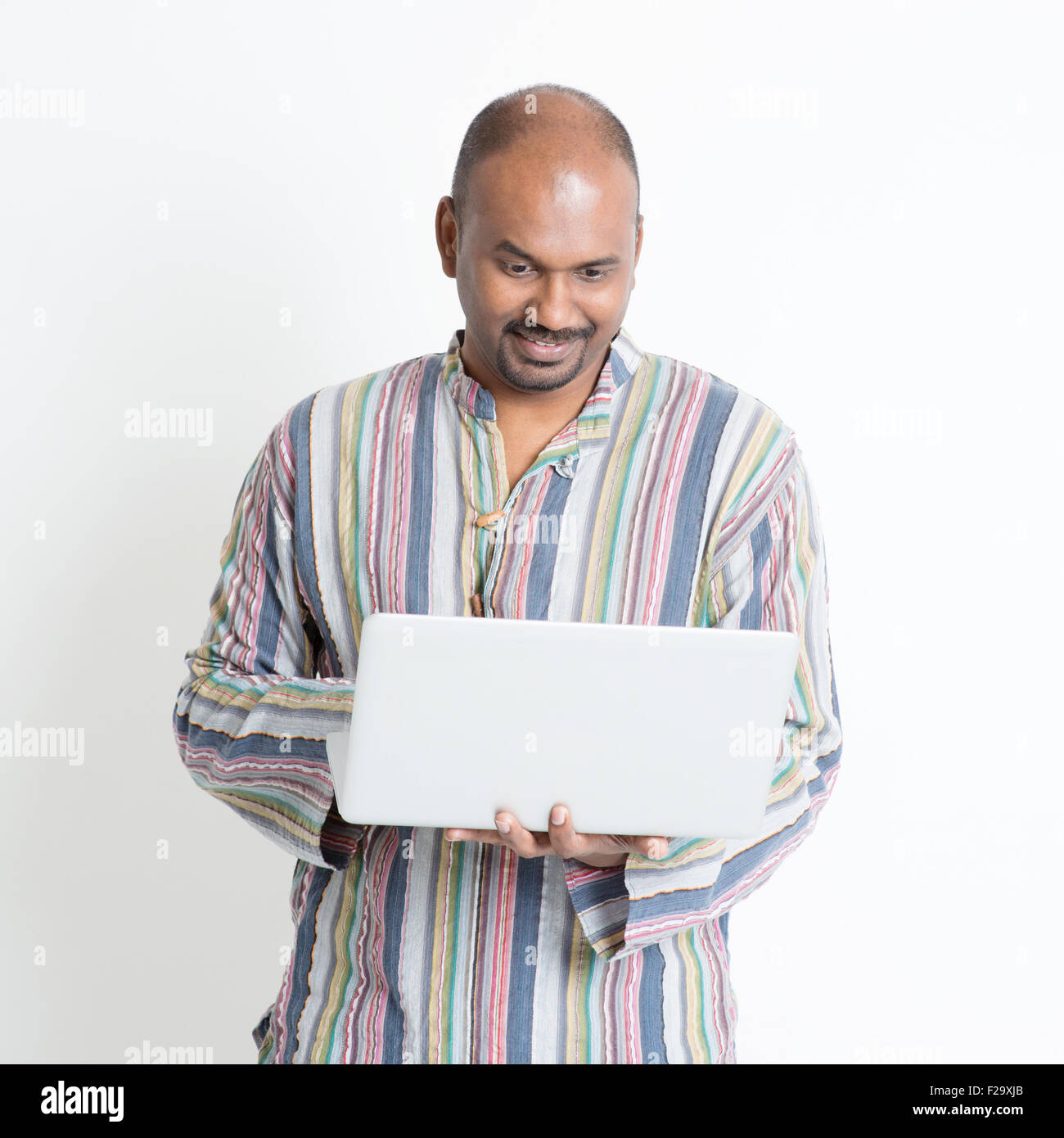 Portrait of mature casual business Indian male using laptop computer, standing on plain background with shadow. Stock Photo