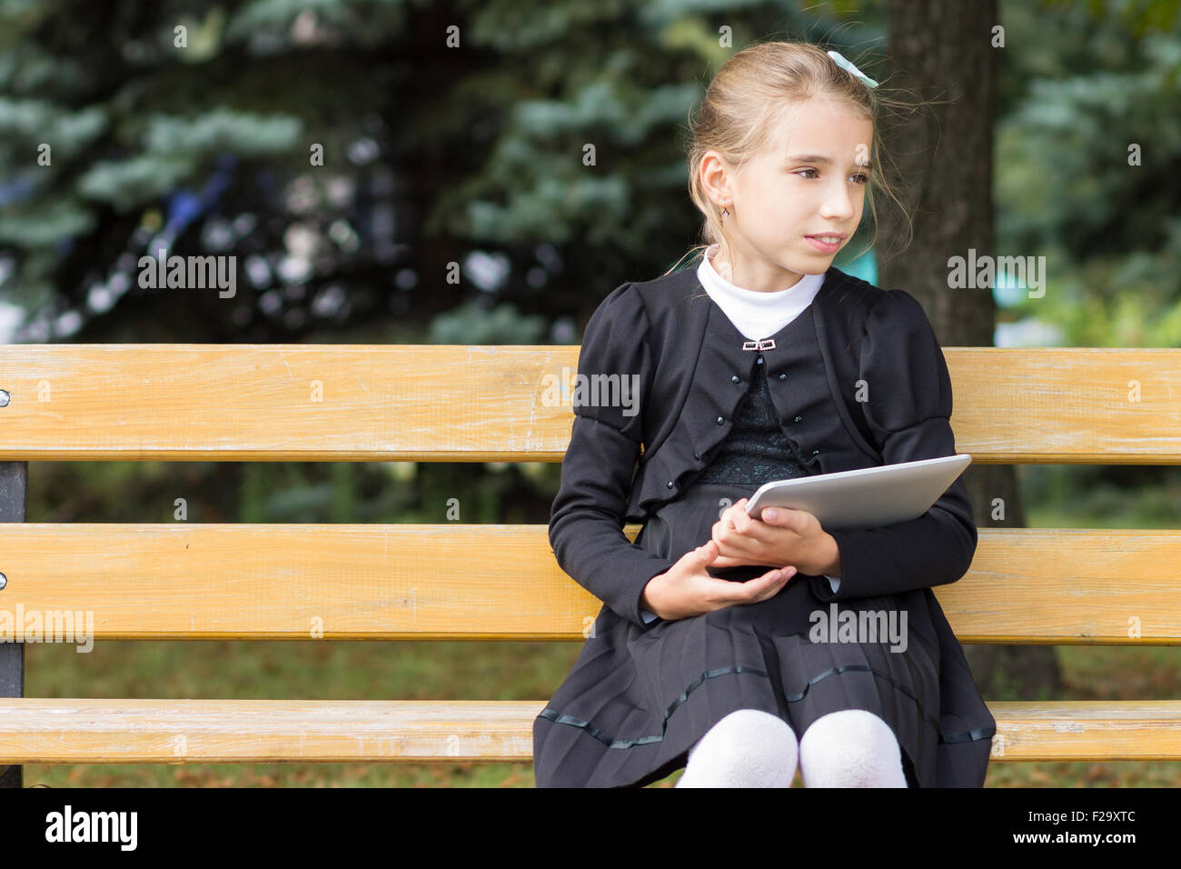 Cute young student girl with tablet sitting in the park. School child resting on the bench outdoors. Copy space area in the left Stock Photo