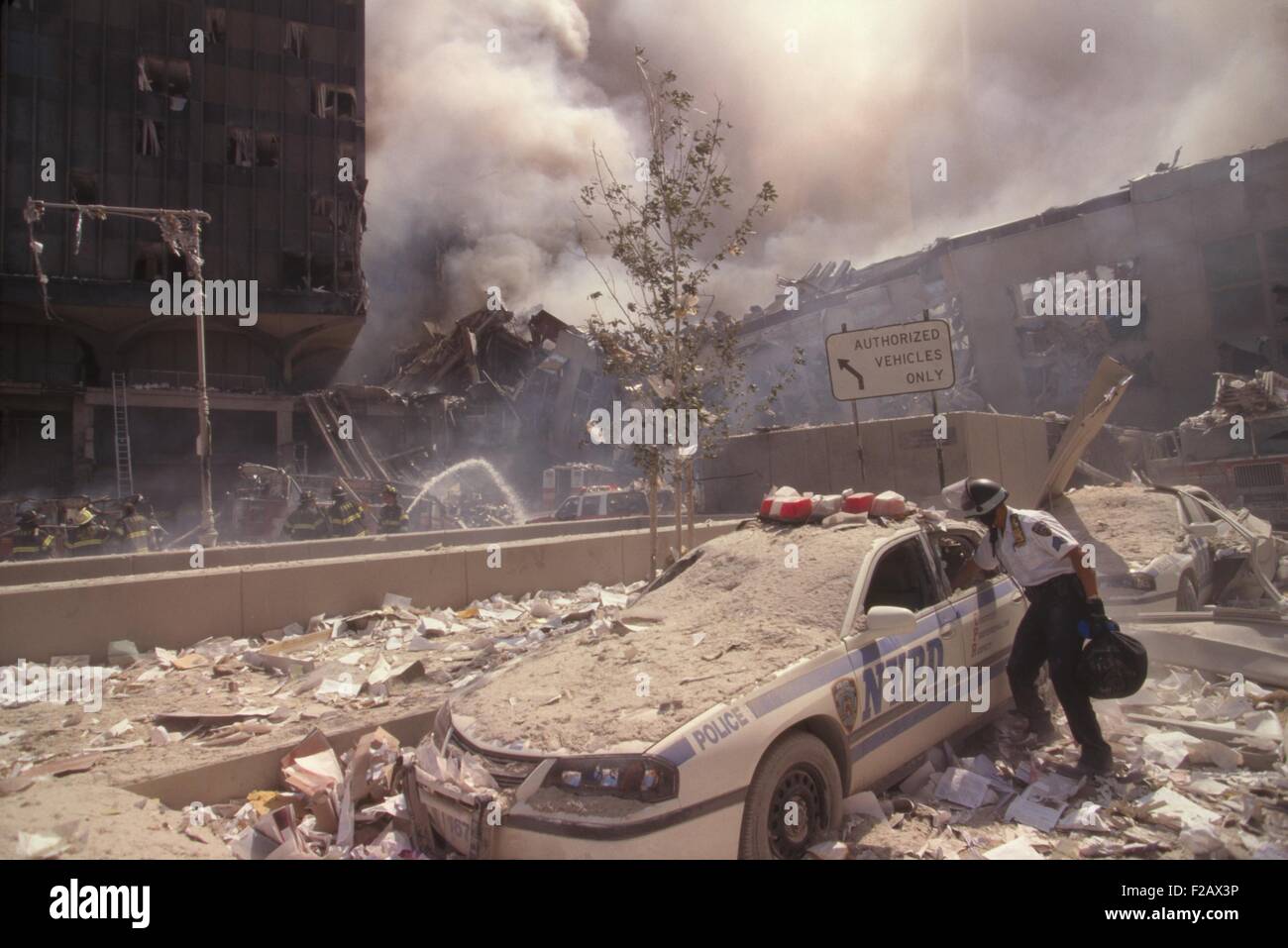Policeman reaching into a debris covered police car after 9-11 terrorist attack in NYC. At left is still standing WTC 6. At right is the destroyed North pedestrian bridge over West Side Highway (West St.). In the background is the burning pile of the collapsed WTC 1 (North Tower). New York City, Sept. 11, 2001. (BSLOC_2015_2_52) Stock Photo