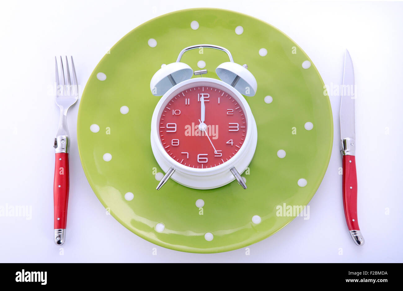 Bright modern table place setting with green polka dot plates and red cutlery on white background with clock set for midnight. Stock Photo