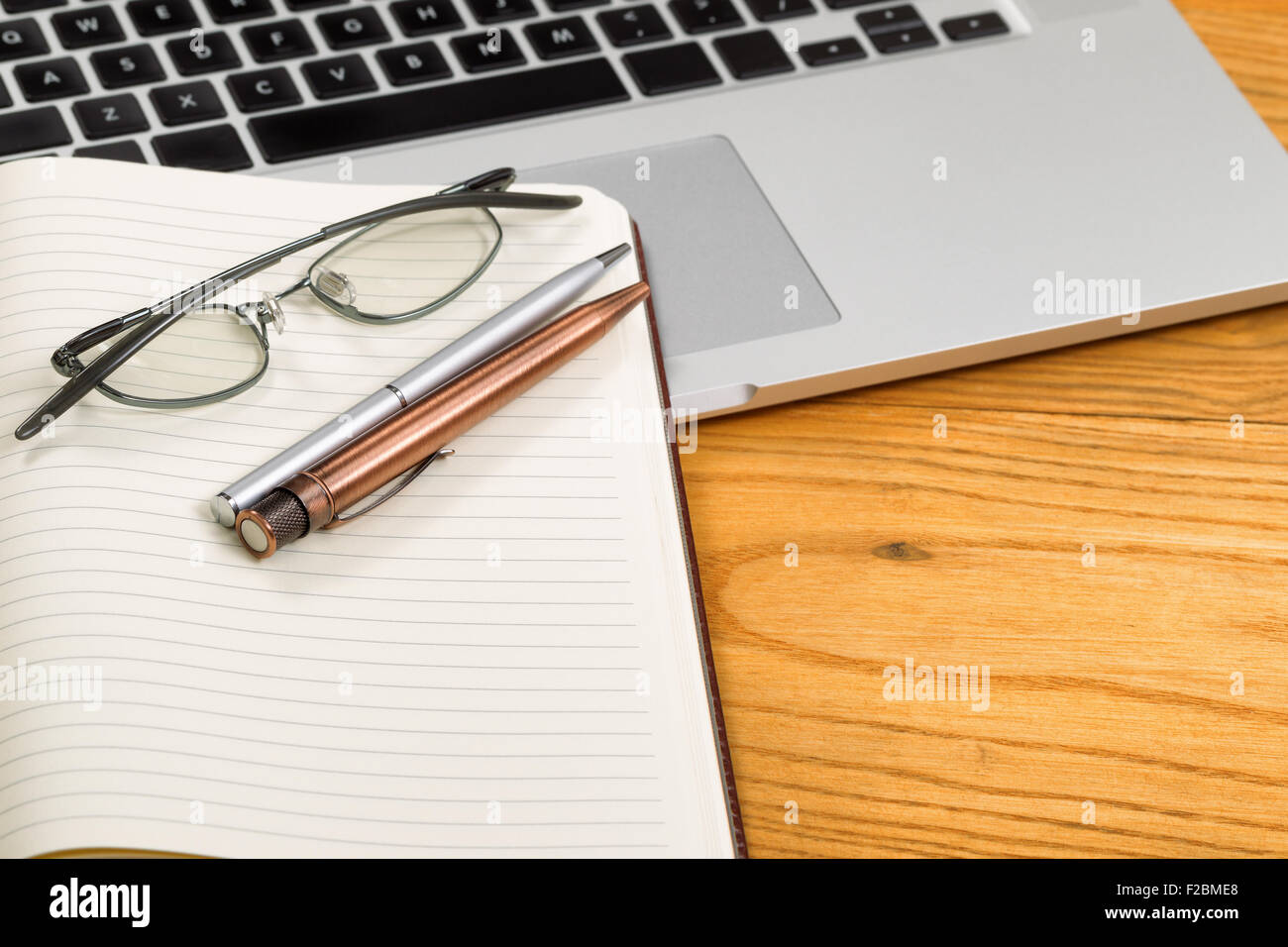 Close up of pens, selective focus on pens, with open blank notebook, reading glasses, and computer in background on desktop. Stock Photo