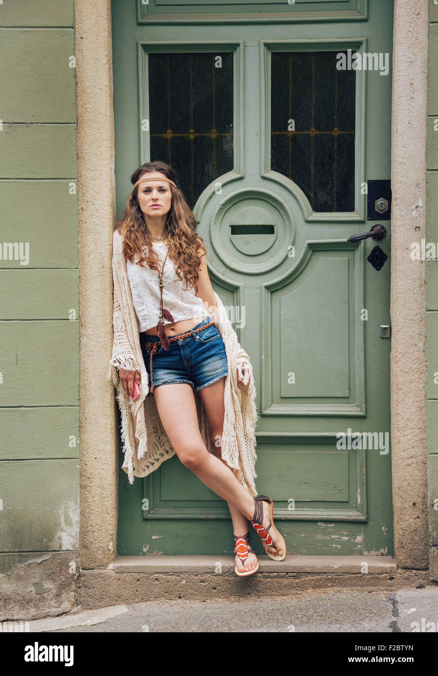 Pensive longhaired hippie woman in bohemian clothes standing  outdoors against wooden door in old town Stock Photo