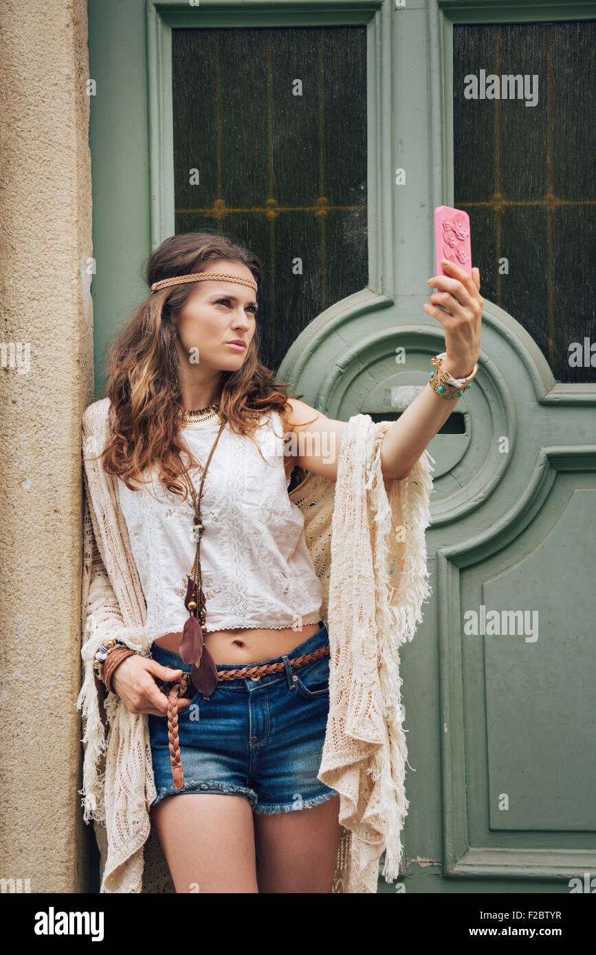 Portrait of hippie woman in boho chic clothes standing outdoors against wooden door in old town and making selfie Stock Photo