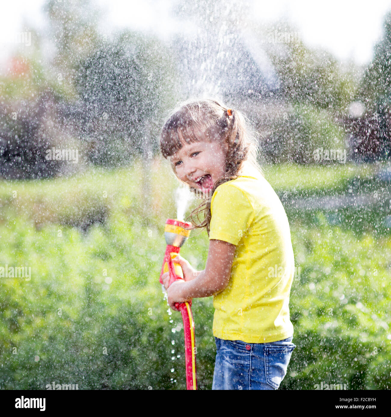 Cheerful kid watering plants from hose spray in garden at backyard of house at sunny summertime Stock Photo