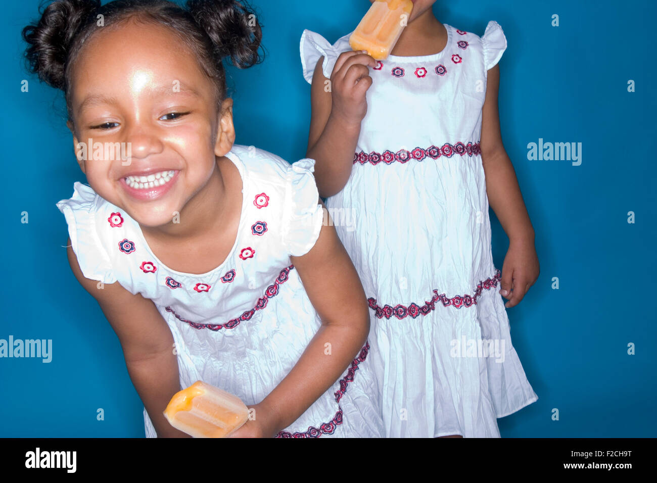 two identical twin girls eat popscicles on blue backgrounds Stock Photo