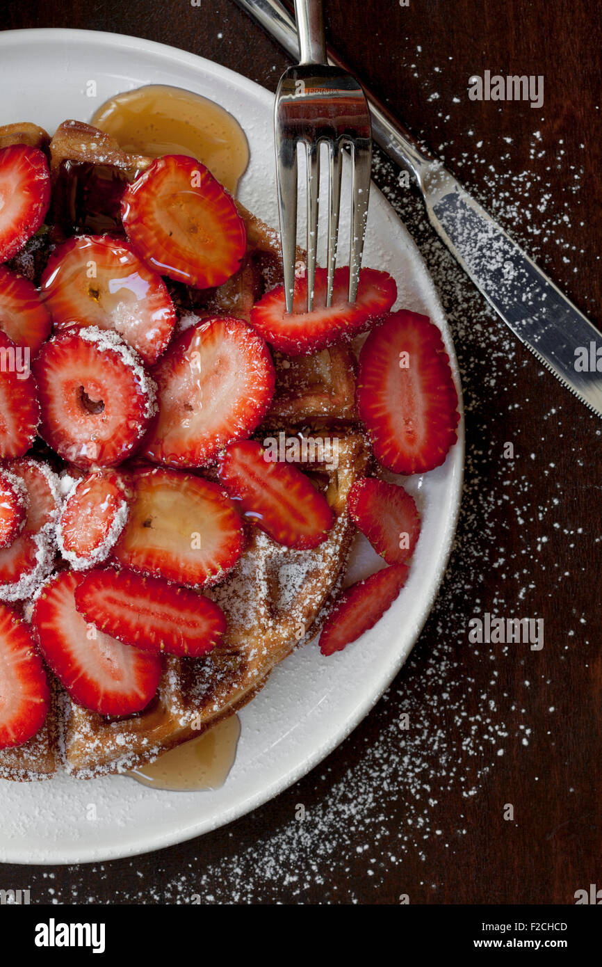 overhead view of a waffle covered with strawberries, powdered sugar and syrup on white plate Stock Photo