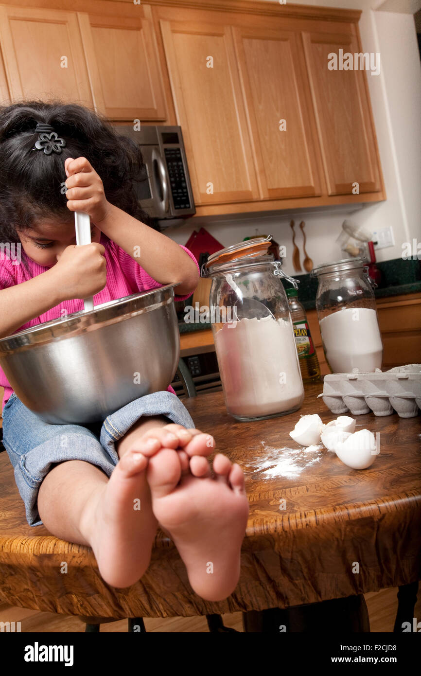 young girl with pink top sits on kitchen counter mixing food in stainless bowl Stock Photo