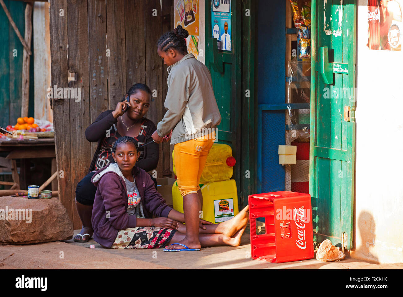Malagasy woman on the phone and girls braiding each other's hair in front of shop in city Ambalavao, Haute Matsiatra, Madagascar Stock Photo