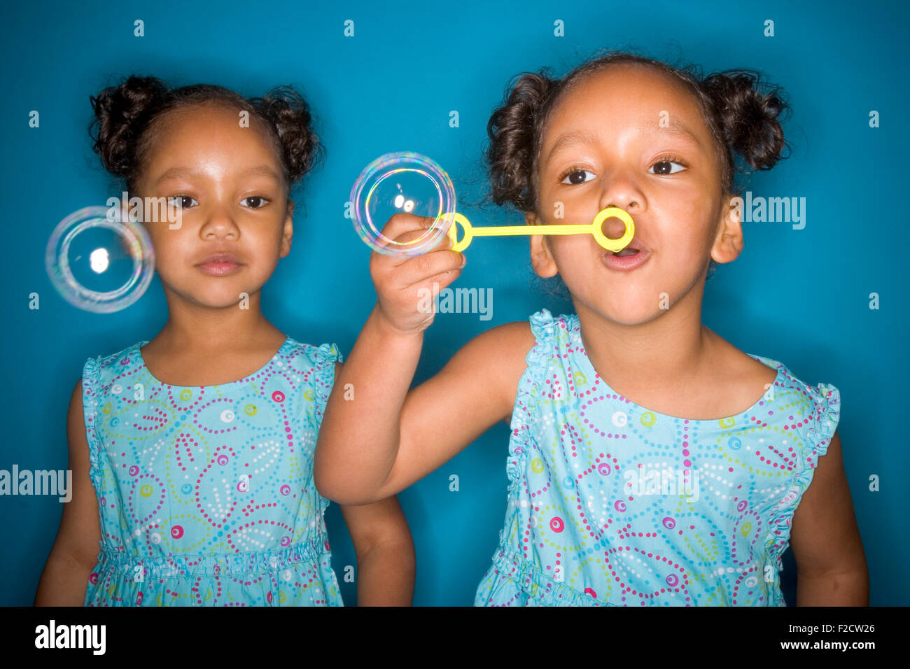 Identical twin black girls wearing matching clothes blow soap bubbles in studio setting Stock Photo