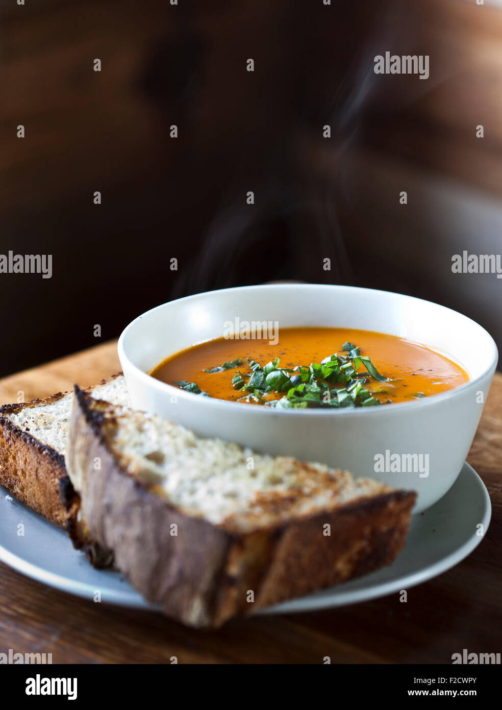 Steaming tomato soup in white bowl with chopped parsley on top and crusty bread on the side in rustic wood out of focus setting Stock Photo