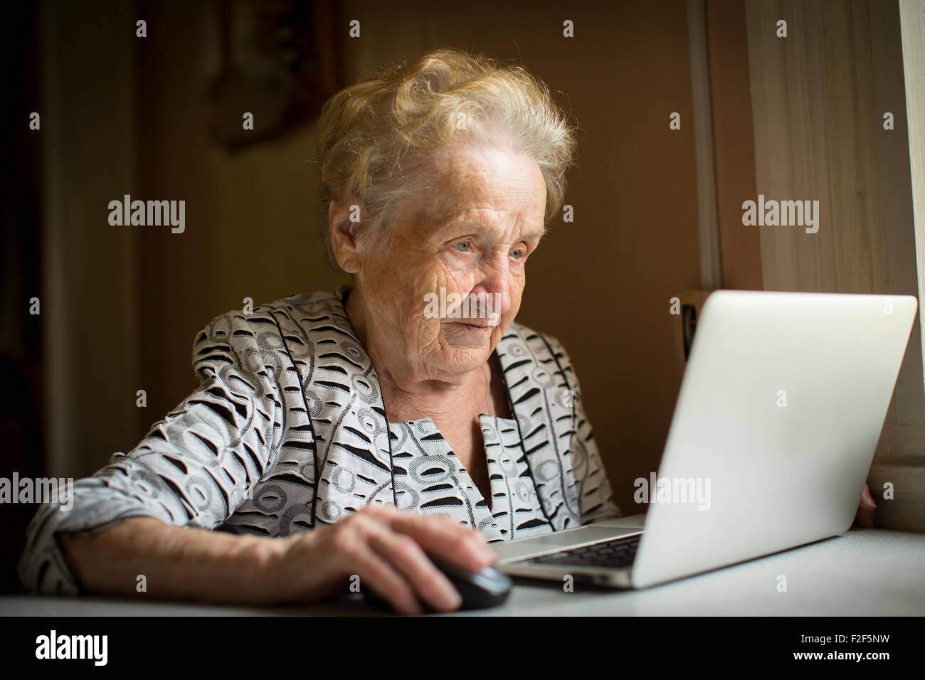Old woman sitting with laptop at table in his house. Stock Photo