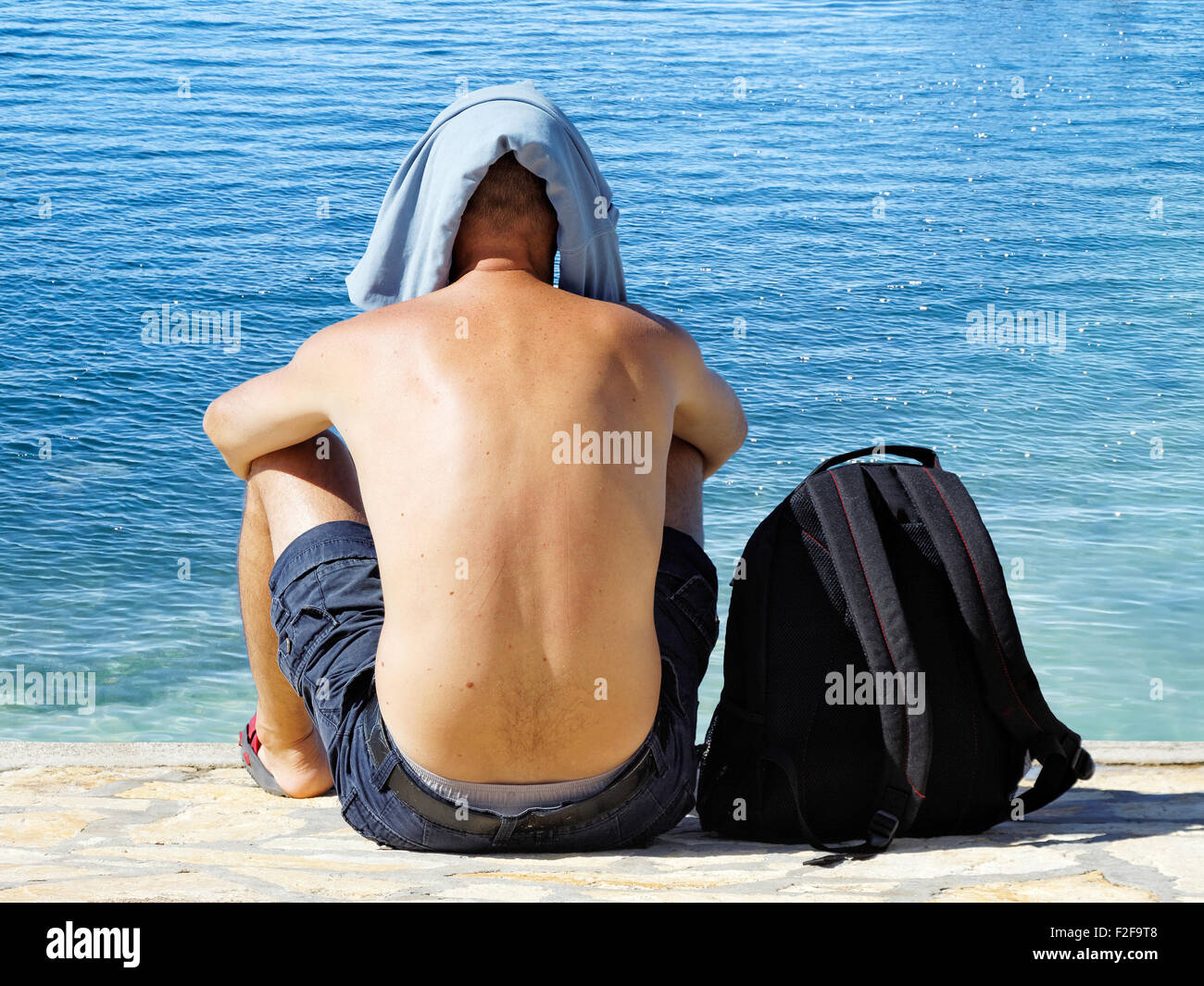 young man sitting on coastline with t-shirt on his head Stock Photo